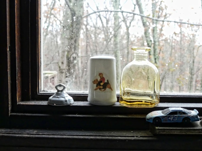 The lid of a tiny, old cache jar. A salt shaker (missing the bottom rubber plug) showing a Norwich Terrier, little glass bottle and a toy car -- on on the narrow sill in the kitchen window